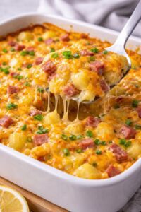 Cheesy egg bake with potatoes, ham, and melted cheese in a casserole dish with a serving lifted